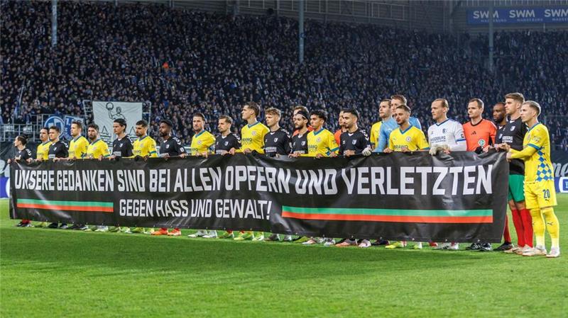 1. FC Magdeburg trotz Patzers Zweitliga-Spitzenreiter Beide Teams stehen während einer Schweigeminute zusammen hinter einem Transparent mit der Aufschrift: „Unsere Gedanken sind mit allen Opfern und Verletzten - gegen Hass und Gewalt!“.