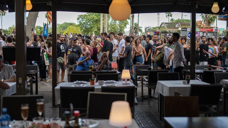 Protest in Barcelona: „Tourists go home“ Ein Mann und eine Frau auf der Terrasse eines Restaurants in Barcelona lassen sich von vorbeiziehenden Demonstranten gegen den Massentourismus offenbar nicht aus der Ruhe bringen.